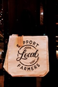 A canvas tote bag hangs against a dark background. The bag features bold, black text that reads 'Support Local Farmers,' accompanied by minimalist decorative elements like wheat stalks. There's a small tag attached to the bag's handle.