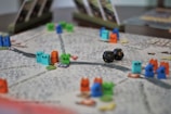A tabletop game board with colorful wooden animal tokens and two black dice. The board is detailed with winding paths and various game cards propped up in the background.