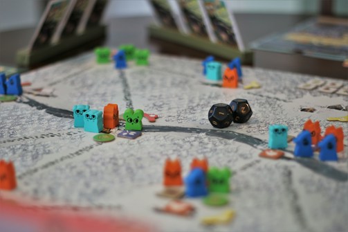 A tabletop game board with colorful wooden animal tokens and two black dice. The board is detailed with winding paths and various game cards propped up in the background.