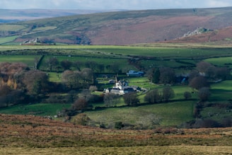 A serene countryside landscape featuring a quaint village nestled amidst green fields and scattered trees. Rolling hills in the background are interspersed with patches of farmland and rocky outcrops. The village consists of several buildings that look like houses and barns.