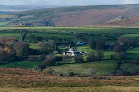 A serene countryside landscape featuring a quaint village nestled amidst green fields and scattered trees. Rolling hills in the background are interspersed with patches of farmland and rocky outcrops. The village consists of several buildings that look like houses and barns.