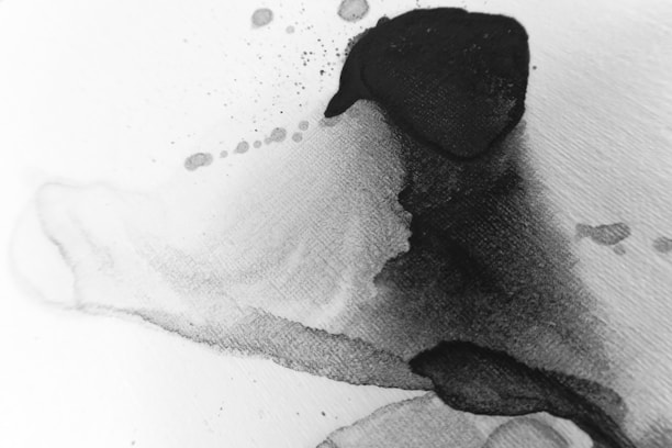 An abstract image featuring fluid, overlapping layers of black ink on a white surface. The ink appears to spread organically with some splatters and gradients visible, creating a dynamic, flowing appearance.