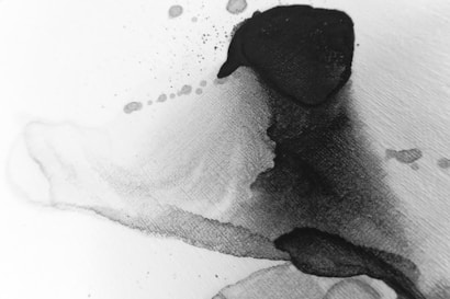 An abstract image featuring fluid, overlapping layers of black ink on a white surface. The ink appears to spread organically with some splatters and gradients visible, creating a dynamic, flowing appearance.