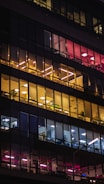 An office building facade at night with illuminated interiors. The windows feature warm yellow and cool white lighting across several floors, showcasing a variety of interior layouts such as open office spaces and individual rooms. Some of the windows reveal plants and office furniture.