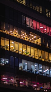 An office building facade at night with illuminated interiors. The windows feature warm yellow and cool white lighting across several floors, showcasing a variety of interior layouts such as open office spaces and individual rooms. Some of the windows reveal plants and office furniture.