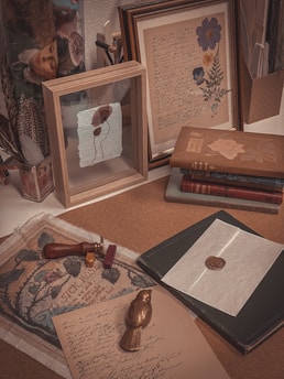 An arrangement of vintage items and writing materials on a desk. There are framed pieces of art, one featuring hand-written text with floral illustrations, and another with a textured paper and dried leaves. Books with ornate covers are stacked, displaying themes of nature. Various writing tools, including a wax seal with two sticks of sealing wax, are placed alongside a cardboard sheet featuring an illustration titled 'The Sick Rose.' The overall aesthetic is warm and nostalgic, suggesting a focus on historical or artistic themes.