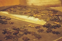 A partially completed jigsaw puzzle is laid out on a surface, with numerous puzzle pieces scattered around it. The puzzle appears to depict a landscape scene, featuring a body of water and trees. The lighting is warm and soft, casting a cozy ambiance over the scene.