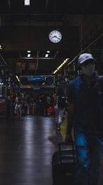 A dimly lit train or bus station interior with a large clock showing 10:10 hanging from the ceiling. Various people are walking and standing around with luggage. The atmosphere appears busy yet subdued, with individuals wearing casual clothing and some carrying bags or suitcases. Overhead lights cast reflections on the dark tiled floor.