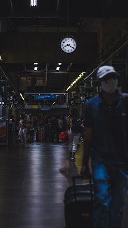 A dimly lit train or bus station interior with a large clock showing 10:10 hanging from the ceiling. Various people are walking and standing around with luggage. The atmosphere appears busy yet subdued, with individuals wearing casual clothing and some carrying bags or suitcases. Overhead lights cast reflections on the dark tiled floor.