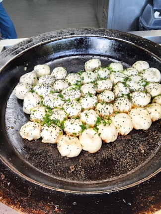 A large black pan is filled with neatly arranged dumplings. The dumplings are topped with black sesame seeds and freshly chopped green herbs.