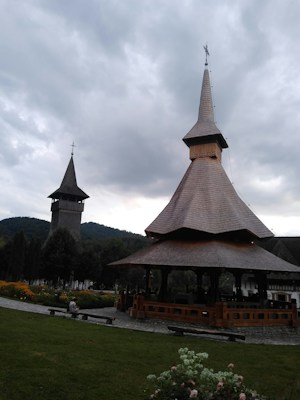A picturesque landscape featuring traditional wooden church structures with tall, pointed spires set against a cloudy sky. The foreground includes well-maintained gardens with colorful flowers and a stone pathway. Lush green hills create a serene backdrop.