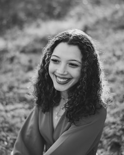 A person with curly hair smiles warmly while looking slightly away from the camera, surrounded by a softly blurred natural background.