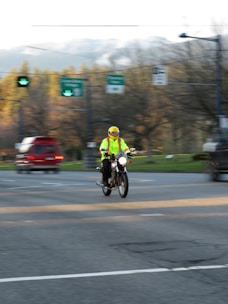 A person wearing a bright yellow safety jacket and helmet is riding a motorcycle. The background features a street intersection with traffic lights and blurred vehicles, indicating motion. Trees and distant hills are visible, suggesting a suburban setting.