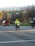 A person wearing a bright yellow safety jacket and helmet is riding a motorcycle. The background features a street intersection with traffic lights and blurred vehicles, indicating motion. Trees and distant hills are visible, suggesting a suburban setting.
