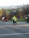 A person wearing a bright yellow safety jacket and helmet is riding a motorcycle. The background features a street intersection with traffic lights and blurred vehicles, indicating motion. Trees and distant hills are visible, suggesting a suburban setting.
