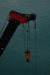 A crane arm with a red and yellow hook hangs over a body of water, with cables suspending the hook. The water is a muted teal color.