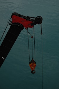 A crane arm with a red and yellow hook hangs over a body of water, with cables suspending the hook. The water is a muted teal color.