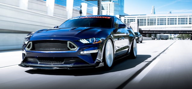 A sleek, dark blue sports car is captured in motion on a city highway. The car features prominent headlights, a bold front grille, and aerodynamic styling. Tall modern buildings and an overpass with large glass windows serve as the backdrop.