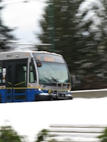 A bus is traveling at speed on a snow-covered road. The surroundings appear blurred due to motion, and evergreen trees are visible in the background. The bus is blue and silver and has a display sign at the front showing its route number.