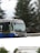 A bus is traveling at speed on a snow-covered road. The surroundings appear blurred due to motion, and evergreen trees are visible in the background. The bus is blue and silver and has a display sign at the front showing its route number.