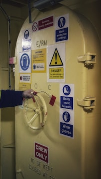 A heavy industrial door is adorned with various safety and warning signs. The door is painted yellow and features a wheel handle, which a person is grasping with their hand. The signs include warnings about wearing protective clothing, boots, and gloves, as well as prohibitions on smoking. There is a notable caution about entering a CO2 protected area. The words 'CREW ONLY' are prominently displayed at the bottom of the door.