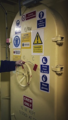 A heavy industrial door is adorned with various safety and warning signs. The door is painted yellow and features a wheel handle, which a person is grasping with their hand. The signs include warnings about wearing protective clothing, boots, and gloves, as well as prohibitions on smoking. There is a notable caution about entering a CO2 protected area. The words 'CREW ONLY' are prominently displayed at the bottom of the door.