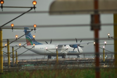 A propeller airplane is situated on a runway, surrounded by several tall, yellow poles equipped with orange lights. The aircraft has a blue, red, and green tail design and belongs to US-Bangla. The atmosphere appears foggy or overcast, suggesting a damp or rainy day.