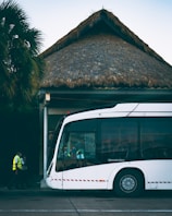A modern white bus is parked outside a building with a thatched roof. Two individuals in high-visibility vests are walking near the bus. Palm trees are visible in the background, adding a tropical atmosphere.