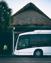 A modern white bus is parked outside a building with a thatched roof. Two individuals in high-visibility vests are walking near the bus. Palm trees are visible in the background, adding a tropical atmosphere.