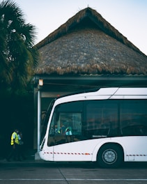 A modern white bus is parked outside a building with a thatched roof. Two individuals in high-visibility vests are walking near the bus. Palm trees are visible in the background, adding a tropical atmosphere.