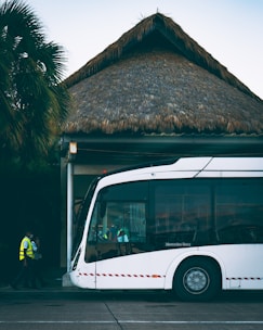 A modern white bus is parked outside a building with a thatched roof. Two individuals in high-visibility vests are walking near the bus. Palm trees are visible in the background, adding a tropical atmosphere.