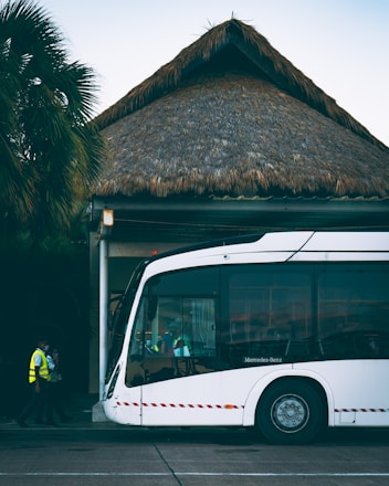 A modern white bus is parked outside a building with a thatched roof. Two individuals in high-visibility vests are walking near the bus. Palm trees are visible in the background, adding a tropical atmosphere.
