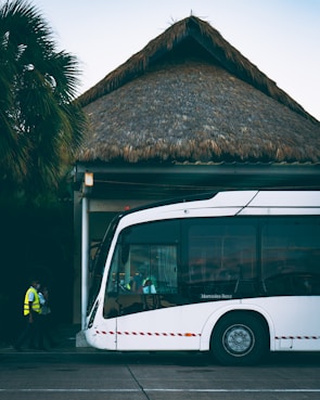 A modern white bus is parked outside a building with a thatched roof. Two individuals in high-visibility vests are walking near the bus. Palm trees are visible in the background, adding a tropical atmosphere.