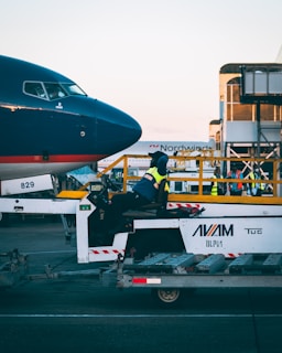A person in a high-visibility vest is seated on a tug vehicle in front of the nose of an airplane on an airport tarmac. The airport setting includes ground support equipment and other workers in the background.