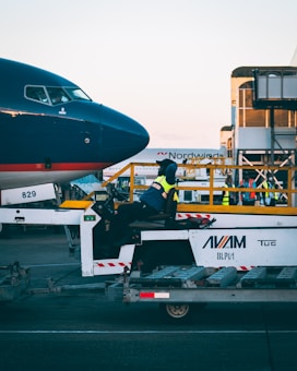 A person in a high-visibility vest is seated on a tug vehicle in front of the nose of an airplane on an airport tarmac. The airport setting includes ground support equipment and other workers in the background.