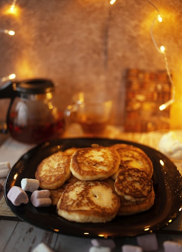 A cozy setup with a plate of golden-brown pancakes accompanied by a few marshmallows. In the background, a glass teapot with tea and a blurred mug are present, set against a warm, softly lit background adorned with string lights.