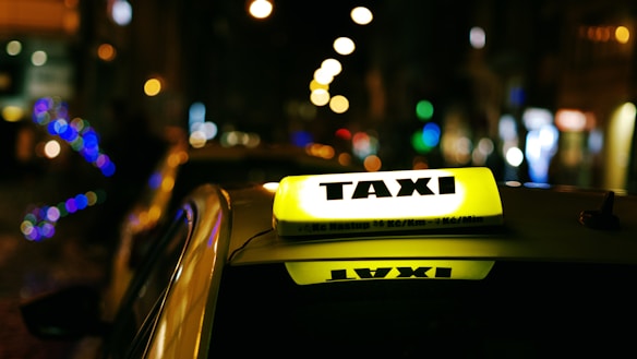 A red taxi with the branding 'Transcab' and a phone number is seen driving along a street. The vehicle is in motion, with a driver visible inside. The surroundings include a textured, gray stone building facade with some surveillance cameras mounted above.
