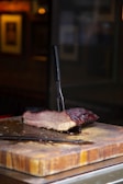 A thick slice of smoked meat rests on a wooden cutting board, with a carving fork lodged into it vertically. The meat has a rich, dark, crusty outer layer and visible juicy layers inside. The background is blurred, highlighting the meat and cutting board in the foreground.