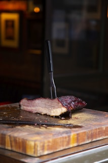 A thick slice of smoked meat rests on a wooden cutting board, with a carving fork lodged into it vertically. The meat has a rich, dark, crusty outer layer and visible juicy layers inside. The background is blurred, highlighting the meat and cutting board in the foreground.