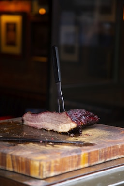 A thick slice of smoked meat rests on a wooden cutting board, with a carving fork lodged into it vertically. The meat has a rich, dark, crusty outer layer and visible juicy layers inside. The background is blurred, highlighting the meat and cutting board in the foreground.