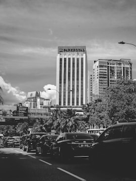 A bustling urban scene with high-rise buildings and heavy traffic. The tall building in the center prominently displays the name 'Parkroyal'. The street is lined with cars, palm trees, and various road signs. The sky is partly cloudy, and shadows are cast across the street.