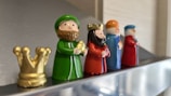 Five small figurines depicting royal or religious figures are lined up on a reflective surface. The figures are dressed in bright colors and hold various objects, with a small golden crown placed next to the first figure. Each figurine has distinctive features and colors.