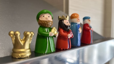 Five small figurines depicting royal or religious figures are lined up on a reflective surface. The figures are dressed in bright colors and hold various objects, with a small golden crown placed next to the first figure. Each figurine has distinctive features and colors.