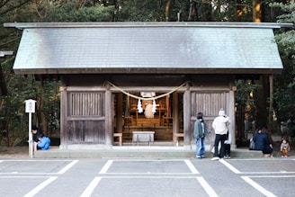 A traditional wooden shrine is positioned in the center, surrounded by people observing or interacting with the space. The small structure features a sloped roof and sliding wooden panels. A rope with white paper streamers is displayed at the entrance. The setting appears serene, with trees providing a lush green backdrop.