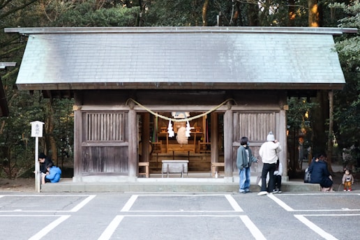 A traditional wooden shrine is positioned in the center, surrounded by people observing or interacting with the space. The small structure features a sloped roof and sliding wooden panels. A rope with white paper streamers is displayed at the entrance. The setting appears serene, with trees providing a lush green backdrop.