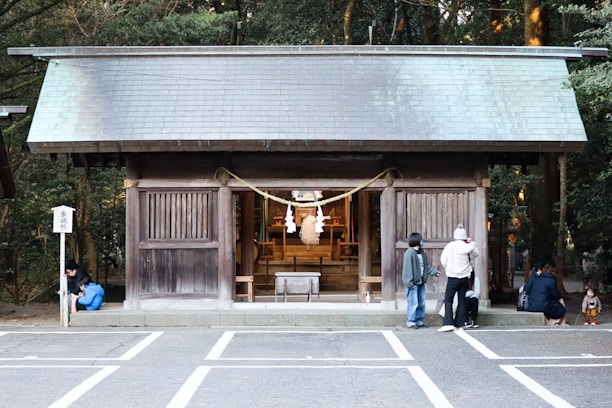 A traditional wooden shrine is positioned in the center, surrounded by people observing or interacting with the space. The small structure features a sloped roof and sliding wooden panels. A rope with white paper streamers is displayed at the entrance. The setting appears serene, with trees providing a lush green backdrop.