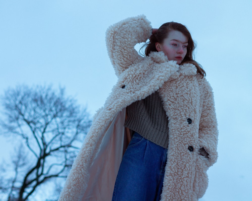 a woman wearing a coat and jeans standing in front of a tree