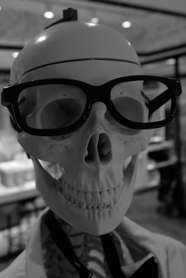 A human skeleton is wearing large, black-framed glasses and is positioned in a well-lit indoor setting with soft background lighting.