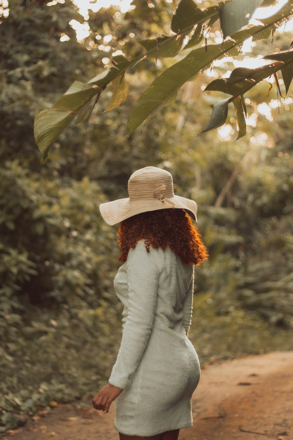 a woman in a white dress and hat walking down a dirt road