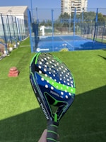 A padel court with blue fencing and a clear blue playing area is shown. A padel racket with a dark blue and green design is held in the foreground. The background includes an artificial grass surface, numbered court signage, and a high-rise building. The setting appears to be an outdoor sports facility.